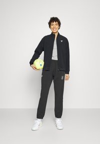 Svart zip-up jacka över en grå tröja, kombinerat med svarta byxor med en logotyp. Håller i en gul fotboll, bär vita sneakers.