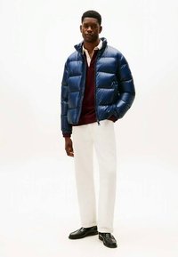 Νεαρός άνδρας φορώντας μπλε φουσκωτό μπουφάν πάνω από ένα μπορντό πουλόβερ και λευκά παντελόνια, στέκεται με το ένα χέρι στην τσέπη και μαύρα παπούτσια.