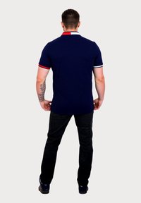 Polo din bumbac de culoare bleumarin, cu guler și mâneci în dungi roșii și albe, asortat cu blugi negri. Designul are o formă ajustată și o textură netedă.