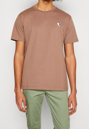 Homme portant un t-shirt marron uni à manches courtes avec un petit logo d'oiseau bleu et blanc sur la poitrine gauche, associé à un pantalon vert clair.