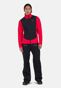Červená a černá vesta s prošívaným předním dílem, s uzávěrem na zip a vysokým límcem. Spárována s černými kalhotami a robustními černými botami.