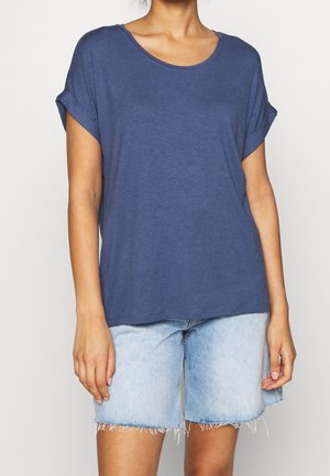 Vrouw draagt een loszittend blauw T-shirt met korte mouwen met opgerolde mouwen en lichtblauwe versleten spijkershorts, staand tegen een witte achtergrond.