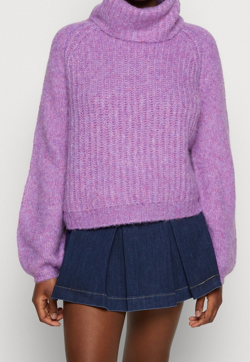 Femme portant un pull à col roulé en tricot violet à manches longues et une mini-jupe plissée bleu foncé.