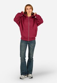 Sweatshirt bordeaux com fecho, bolso frontal, combinado com jeans flare de ganga e ténis cinzentos. Apresenta um capuz espaçoso e um corte descontraído.