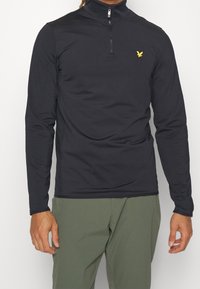 Svart långärmad zip-up topp i ett mjukt och flexibelt material. Den har en hög krage och en liten gul logotyp på vänster bröst.