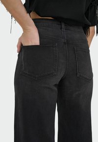 Jeans en denim noir avec une taille cintrée et des jambes larges. Comprend deux poches plaquées à l'arrière, des coutures visibles et une texture lisse.