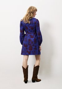 Lila Kleid mit einem gemusterten Design in dunklen roten Formen und floralen Motiven, kombiniert mit braunen Wildleder-Cowboy-Stiefeln.