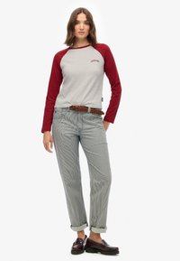 Kobieta w szarej i czerwonej koszuli z długim rękawem, w paskowanych, podwiniętych spodniach oraz brązowym plecionym pasku, w ciemnobrązowych mokasynach, stojąca na białym tle.