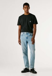 Černé bavlněné tričko s kapsou na hrudi, světle modré džínové kalhoty a černé kožené boty se šňůrkami. Model stojí uprostřed na bílém pozadí.