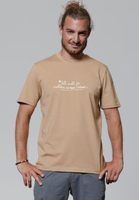 Mann mit zurückgebundenem Haar und Bart, der ein beiges T-Shirt mit deutschem Text trägt und vor einem schlichten grauen Hintergrund steht.