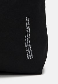 Svart väska tillverkad av återvunnen bomull med en texturerad yta. Har vittryckt text på ena sidan. Enkel design, inga ytterligare mönster.