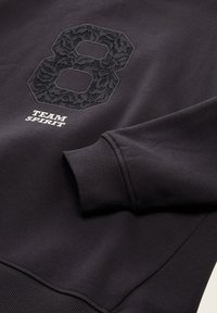 Sweatshirt noir avec un numéro "8" en dentelle texturée et le texte "TEAM SPIRIT" en blanc. Manches côtelées et texture de tissu lisse.
