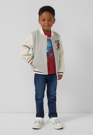 Jongen staat, draagt een grijs-witte varsityjack, een rood graphic t-shirt, blauwe spijkerbroek en witte sneakers tegen een effen achtergrond.