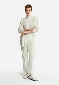 Cremefarvet ribstrikket sweater med pufærmer, parret med hvide skræddersyede bukser og lyse loafers, sat mod en hvid baggrund.