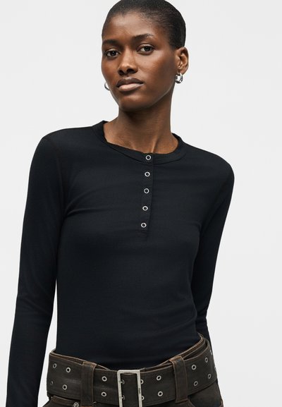 Γυναίκα που φοράει μαύρο μακρυμάνικο ριπ τοπ με κουμπιά σφικτήρα και φαρδιά μαύρη ζώνη με μεταλλικές τρυπούλες και αγκράφα.