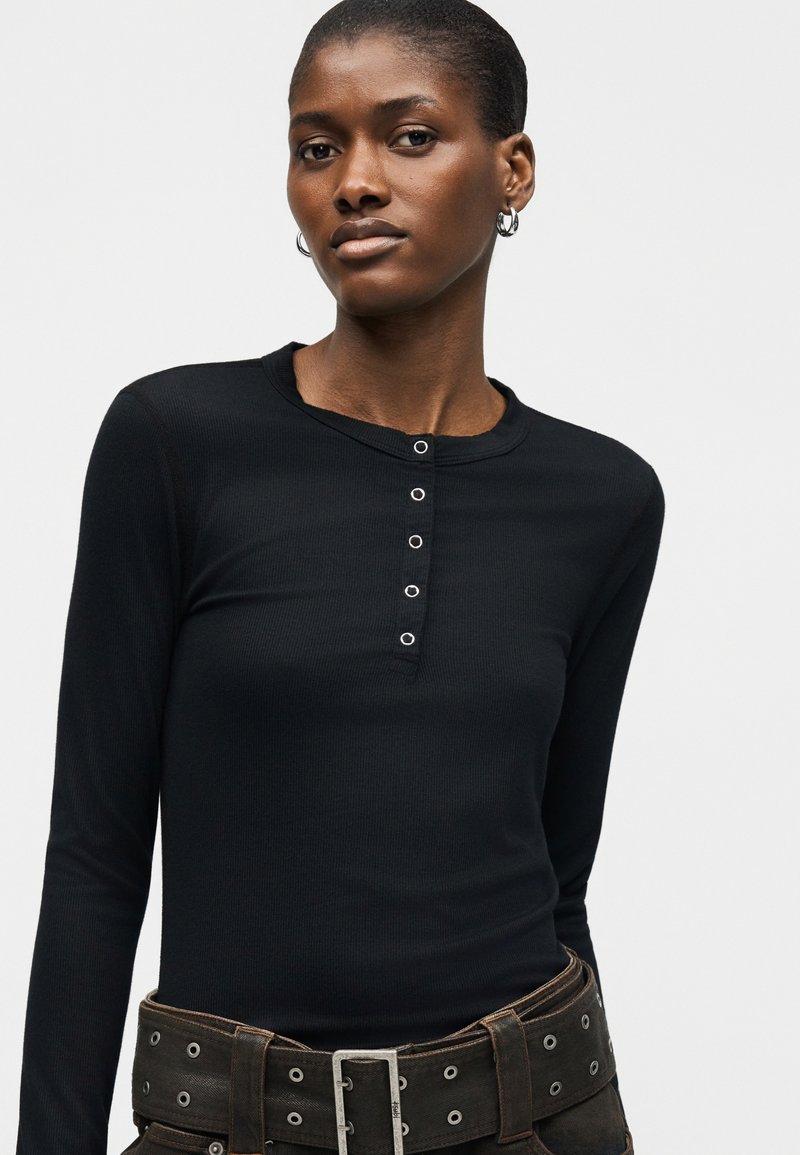 Woman wearing a black long-sleeve ribbed top with snap buttons and a wide black belt with metal eyelets and buckle.