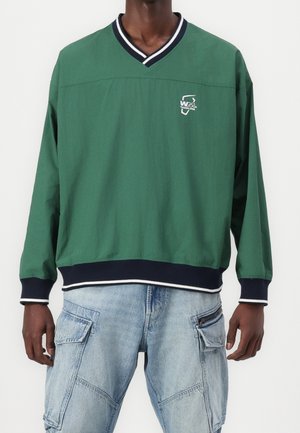 Person wearing a green long-sleeve pullover with navy and white striped V-neck and cuffs, paired with light blue cargo jeans.