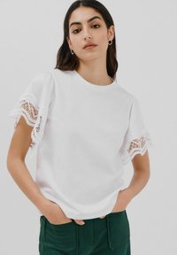 Hvid bomulds T-shirt med korte ærmer med blondekant. Stoffet er glat og har en rund halsudskæring, stylet med mørkegrønne bukser.