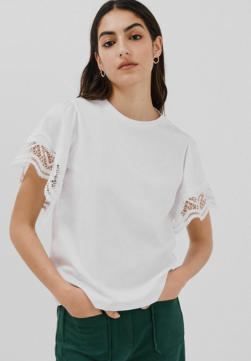 Hvid bomulds T-shirt med korte ærmer med blondekant. Stoffet er glat og har en rund halsudskæring, stylet med mørkegrønne bukser.