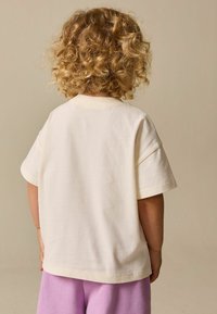 Krémově zbarvené bavlněné tričko s volným střihem, krátkými rukávy a jednoduchým designem, nošené s fialovými plyšovými kalhotami. Zobrazen pohled zezadu.