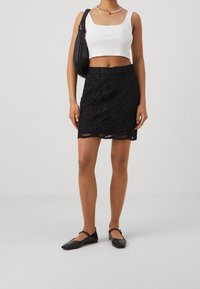 Kvinna klädd i en vit kortärmad tank top, svart spets mini kjol, svarta Mary Jane skor, pärlhalsband och bär en svart axelväska.