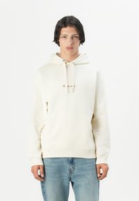 Crèmekleurige hoodie met een voorzak in kangoeroestijl, een capuchon met trekkoord en subtiele geborduurde tekst in bruin. Gecombineerd met lichtblauwe jeans.