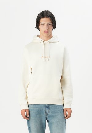 Cream hoodie with a front kangaroo pocket, drawstring hood, and subtle embroidered text in brown. Paired with light blue jeans.