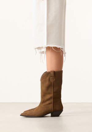 Benen die witte cropped broeken dragen met rafelige zomen en bruine suède cowboylaarzen staan op een lichte vloer.