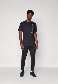 Muž v černém tričku značky Tottenham Hotspur a Nike, černých kalhotách Nike a černých botách, stojící proti jednobarevnému světlému šedému pozadí.