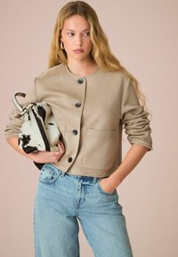 Veste beige raccourcie avec un col rond, des boutons noirs et deux poches avant. Associée à un jean taille haute bleu clair et un sac à motifs.