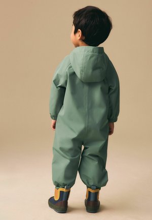 Laps seisab seljaga vaataja poole, seljas roheline kapuutsiga vihmamantel ja jalas mitmevärvilised kummisaapad, taust neutraalne.