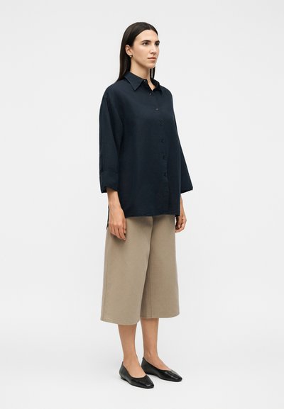 Γυναίκα όρθια, φοράει ένα φαρδύ μαύρο πουκάμισο με κουμπιά και μακριά μανίκια, μπεζ φαρδιά καλοκαιρινά παντελόνια και μαύρα φλατ παπούτσια μπροστά σε μονόχρωμο φόντο.