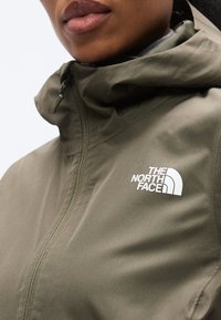 Αδιάβροχο μπουφάν σε ελιά πράσινο με ψηλό γιακά, εμπρόσθιο φερμουάρ και λευκό λογότυπο στο αριστερό μέρος του στήθους. Ομαλή υφή υφάσματος.