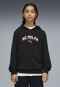 Sweat à capuche noir avec le logo "AC MILAN" en blanc et rouge. Comprend une poche avant et des poignets côtelés. Porté par-dessus une chemise blanche.