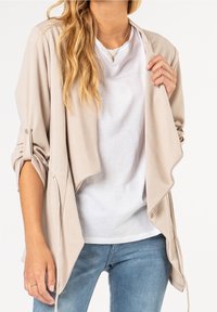 Beiger, leichter Cardigan mit vorne drapiertem Schnitt, hochgekrempelten Ärmeln und dezentem Glanz, getragen über einem weißen T-Shirt und blauen Jeans.