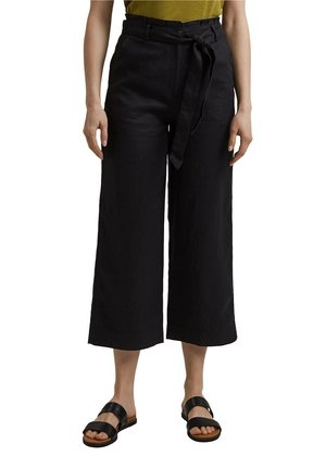 Pantalons larges noirs à taille ceinturée, en tissu lisse. Portés avec des sandales noires et un haut vert.