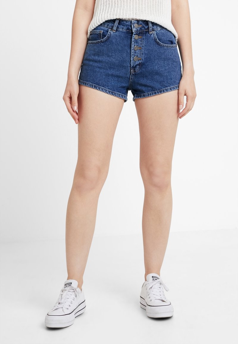 Džínové šortky s vysokým pasem, zapínáním na pět knoflíků a roztrhaným lemem. Tmavě modrá barva s mírným vyblednutím. Kombinované s bílými nízkými teniskami.