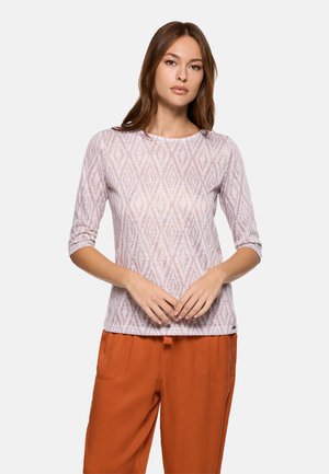 Joven con cabello largo y castaño, que lleva una blusa ligera de manga tres cuartos con un patrón y pantalones sueltos naranjas, de pie con las manos entrelazadas al frente.