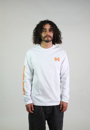 Mann mit lockigem Haar, trägt ein weißes Langarmhemd mit orangem Text auf der Brust und am linken Ärmel, steht vor einem schlichten hellen Hintergrund.