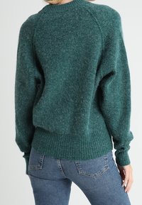 Vihreä neulottu villapaita, jossa on tekstuuri, ribbaattu helma ja hihansuut sekä raglan-hihat. Yhdistetty sinisiin farkkuihin, jotka näyttävät rennolta istuvalta.