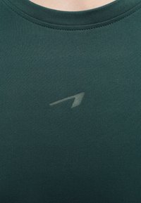 Κοντινή λήψη σκοτεινού πράσινου αθλητικού πουκαμίσου με μικρό, ανοιχτό γκρι λοξό βελάκι λογότυπο κεντραρισμένο κάτω από το λαιμόκοψη.