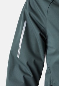 Manche de veste imperméable verte avec un design épuré, une texture lisse et une bande réfléchissante blanche avec le logo "SWEDEMOUNT".