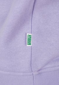 Tissu tricoté lavande avec une petite étiquette blanche cousue dans la couture, affichant le texte vert « URBAN » en position verticale.