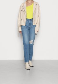Chaqueta de cuero beige con cremalleras, top amarillo brillante de un solo hombro, jeans azul claro desgastados y botines crema con detalles en plata.