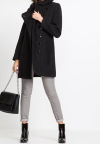 Svart kappa med hög krage, knapppdetalj och långa ärmar; kombinerad med grå, slim fit-jeans och blanka svarta ankelstövlar.