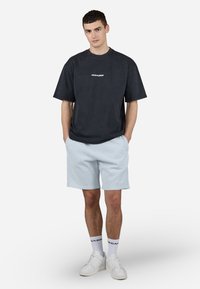 Svart bomull T-skjorte med "PEGADOR" tekst i hvitt, sammen med lyseblå ribbede shorts. Modellen har på seg hvite joggesko og ankelsokker.