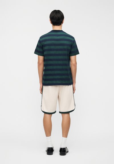 Μπλε ναυτικό και σκούρο πράσινο ριγέ κοντομάνικο μπλουζάκι σε συνδυασμό με εκρού σορτς με σκούρα πράσινη διακόσμηση. Μαύρα παπούτσια με λευκές κάλτσες.