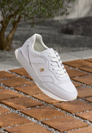 Biały tenisówka ze sznurówkami i złotym logo, ustawiona nad drewnianą powierzchnią z małymi szarymi kamieniami między płytkami.