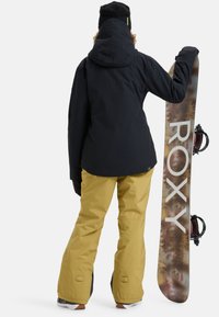 Black snow jacket with a hood, paired with yellow snow pants. A snowboard with a nature-themed design and logo rests against the figure.