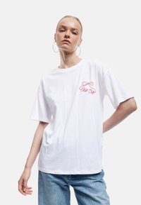 Hvid bomulds T-shirt med korte ærmer, med en rød broderet sløjfe og teksten "Chill Out" på venstre bryst. Løs pasform.